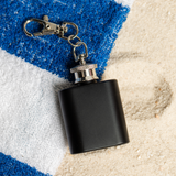 Matte black 1oz keychain flask with clip on a striped towel and sand, ready for engraving