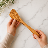 Hands holding a 12 inch olive wood spatula with an angled edge over a marble counter