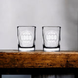 Two custom 2.2 oz shot glasses engraved with a Your Logo Here sample on a wooden shelf