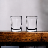 Two plain 2.2 oz square shot glasses on a wooden shelf, ready for custom engraving