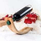 Handmade olive wood wine bottle holder with a curved balancing base holding a red wine bottle beside gifts