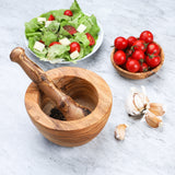 Plain olive wood mortar and pestle with peppercorns inside, next to a salad, tomatoes and garlic