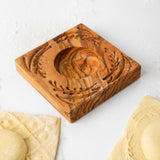 Close-up of an olive wood ravioli mold with a carved leaf wreath pattern beside fresh ravioli