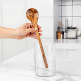 Hand holding a long olive wood cooking spoon above a glass holder in a kitchen