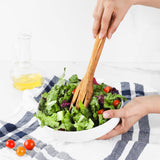 Hands tossing a bowl of salad with a long olive wood fork, olive oil bottle in the background