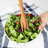 Hand serving salad with an olive wood fork from a white bowl on a striped towel
