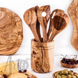 Olive wood utensil holder filled with spoons and spatulas beside cherries, olives and a cutting board