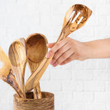 Hand taking a slotted olive wood spatula from a utensil crock filled with spoons
