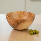 Round olive wood bowl from the side next to a few green olives on a pine counter