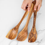 Hand holding three handmade olive wood corner spoons with angled tips over white marble