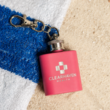 Pink 1oz keychain flask engraved with a sample health company logo on a striped beach towel